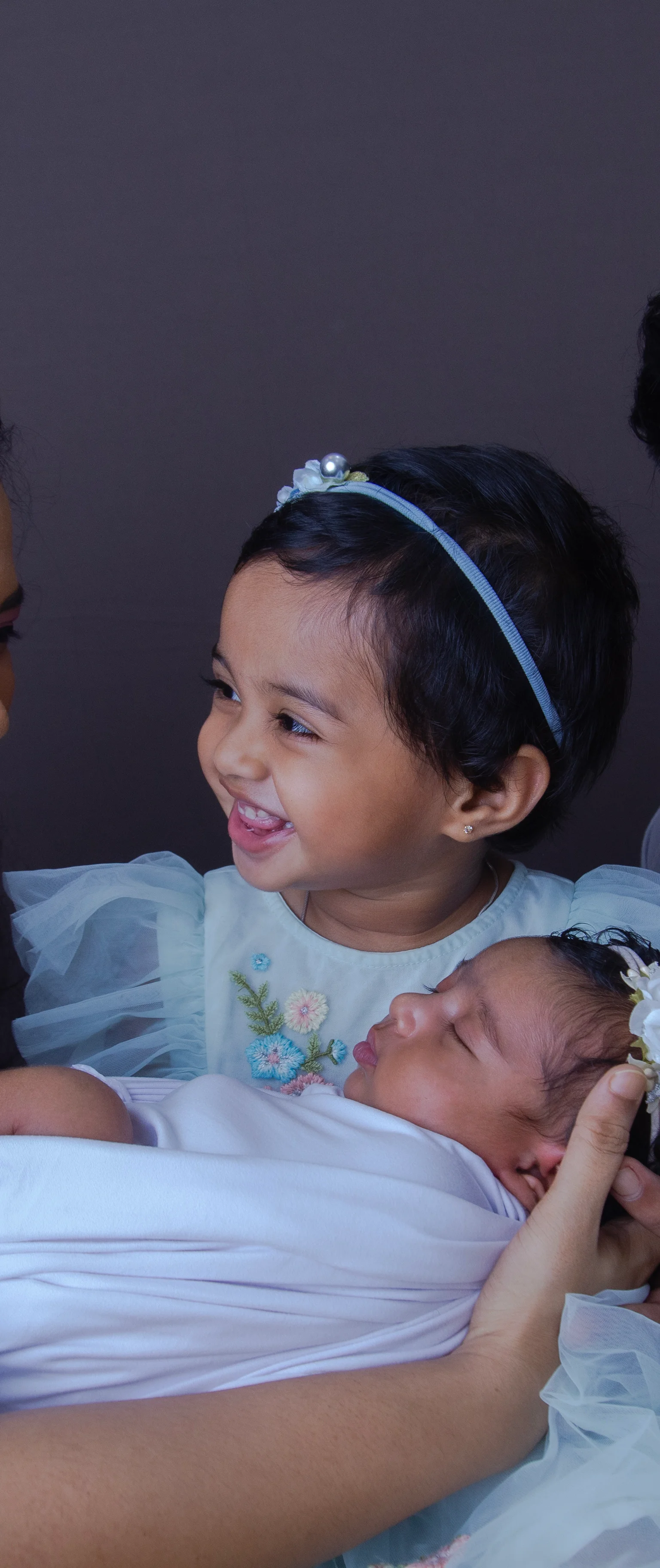 A studio portrait of a new mother, father, their newborn daughter along with the newborn baby's sibling. The portrait is extremely cute and they look very happy.